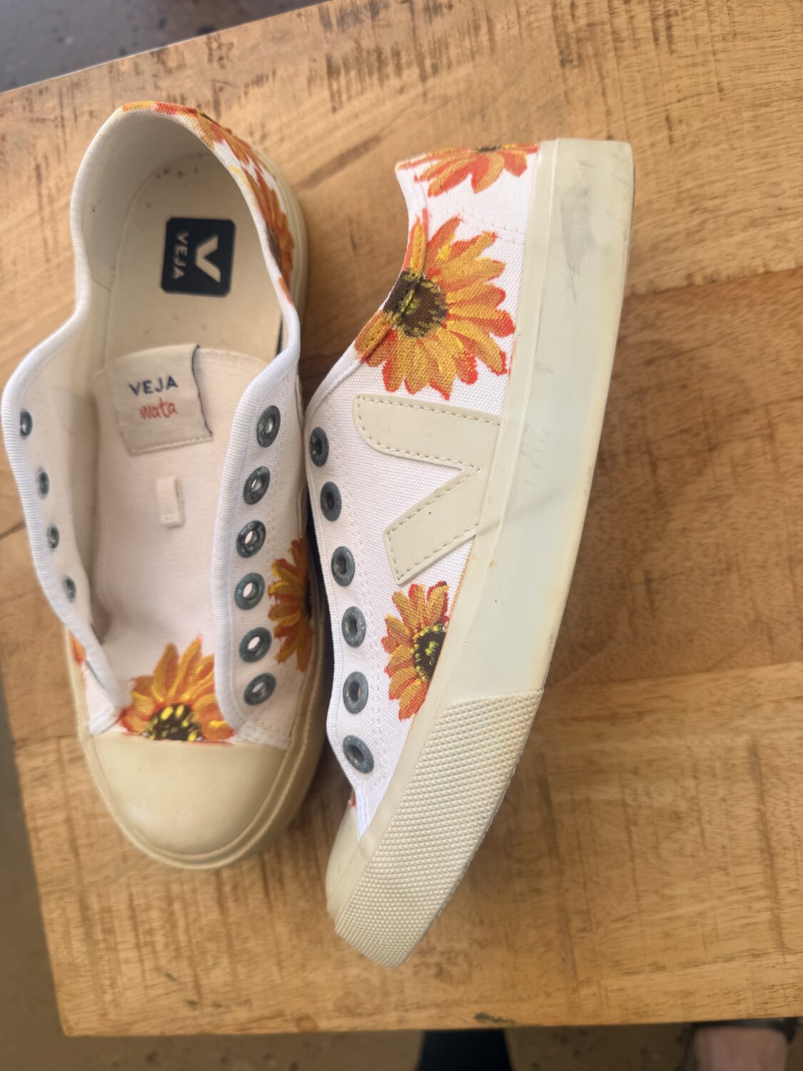 Pair of white canvas sneakers with bright orange sunflower prints, placed on a wooden table, Veja branding visible inside