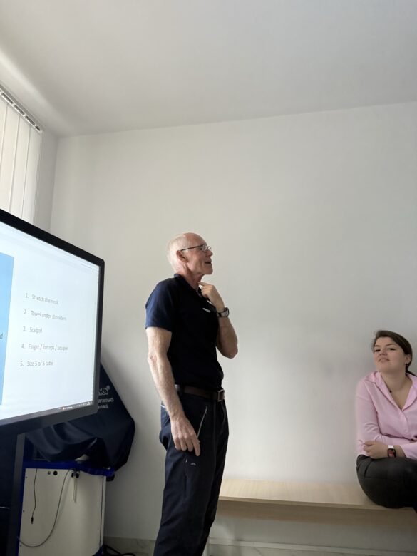 Older man in a dark polo standing and speaking to a classroom, with a young woman seated on a bench beside a large monitor displaying a slide.