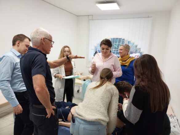 Group of students and instructors gathered around a patient on a hospital bed for a clinical training demonstration.",