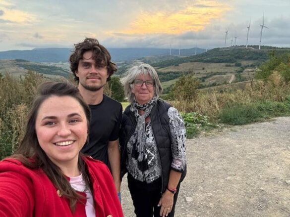 Three people pose for a selfie on a dirt trail with rolling hills and wind turbines in the distance.