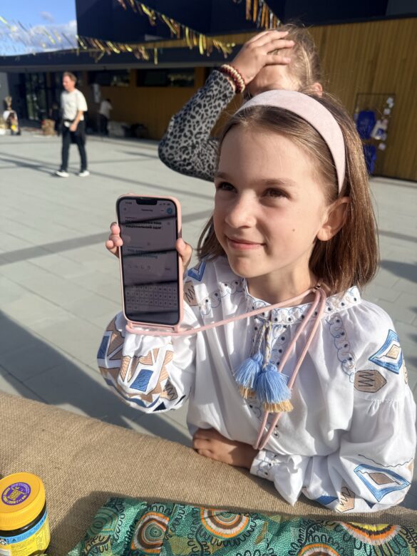 IMG_6440 Smiling young girl with a pink headband holding a pink smartphone outdoors in daylight, showing the screen to the camera.
