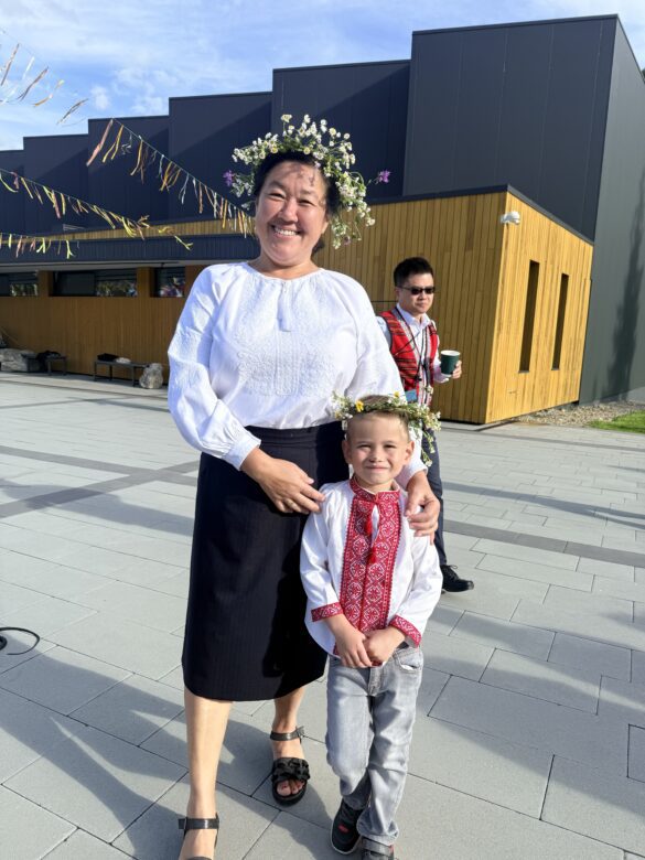 IMG_6442 Woman and child in traditional embroidered clothing with flower crowns posing outdoors in front of a modern building; both smiling.