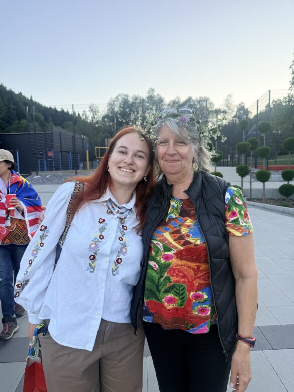 IMG_6469 Two women posing together outdoors, smiling, one wearing a flower crown and a colorful top beside a younger woman in an embroidered white blouse.
