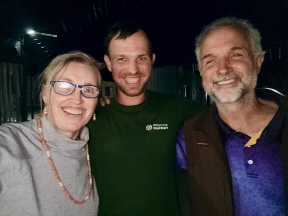 IMG_6486 Three friends pose for a selfie at night, smiling at the camera: woman with glasses on the left, man in green shirt center, man in purple shirt and brown vest on the right.
