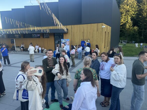 IMG_6523 Group of people in traditional embroidered shirts posing and chatting on a paved plaza in front of a modern wood-paneled building with decorative pennant banners overhead, some taking a selfie nearby.