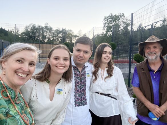 IMG_6524 Five people pose for a selfie outdoors, smiling, wearing traditional embroidered white blouses and a patterned shirt, with a fenced sports area in the background.