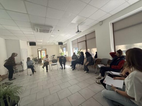 Group of adults seated in a circle in a bright, tiled room; a facilitator stands at the left side of the circle.