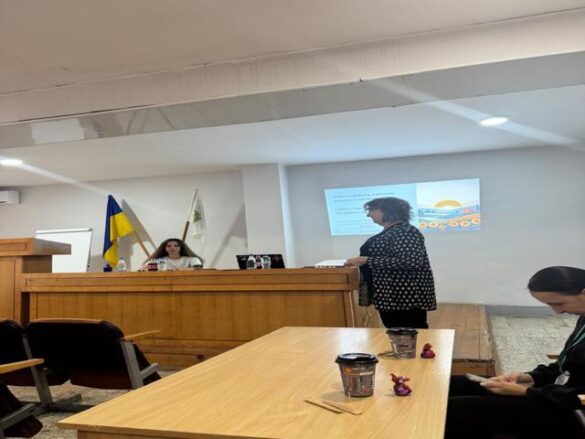 Panel of speakers seated at a wooden dais in a conference room, Ukrainian flag and another flag behind them, presenter standing to the right with a projected slide on the screen