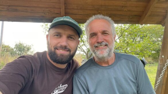 Two men smiling for a selfie under a wooden shelter, the younger man wearing a green cap and brown shirt, the older man with a gray beard and blue shirt, outdoors on a sunny day.