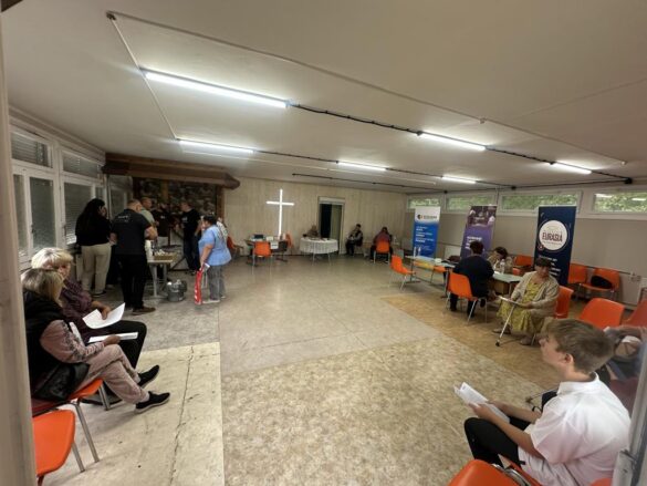 People waiting and being assisted in a well-lit community room; orange chairs line the space, banners stand along the walls, and a cross is visible at the back.