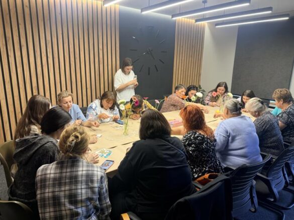 Group of adults around a long craft table, working on projects as an instructor in a white shirt stands at the head of the table with flower vases in the center.