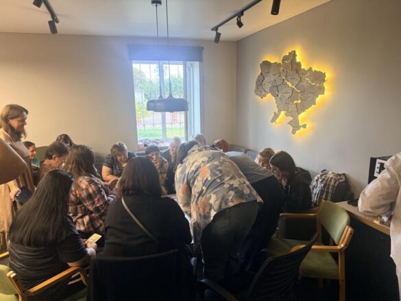 A group of adults gathered around a table in a meeting room, leaning in to work on a shared task, with a window behind them and a lit wall map on the right.