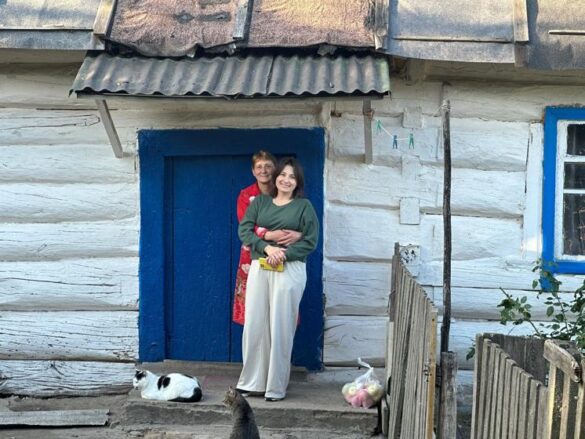 Two women stand arm-in-arm in front of a white cottage with a blue door; a black-and-white cat rests on the steps nearby.