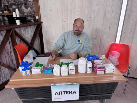 Man with glasses sits at a table filled with medication bottles and boxes, signing papers; 'АПТЕКА' sign on the front of the desk.