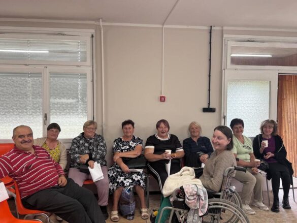Group of about ten adults seated along a hallway waiting area, one person in a wheelchair at the front, all smiling or chatting.