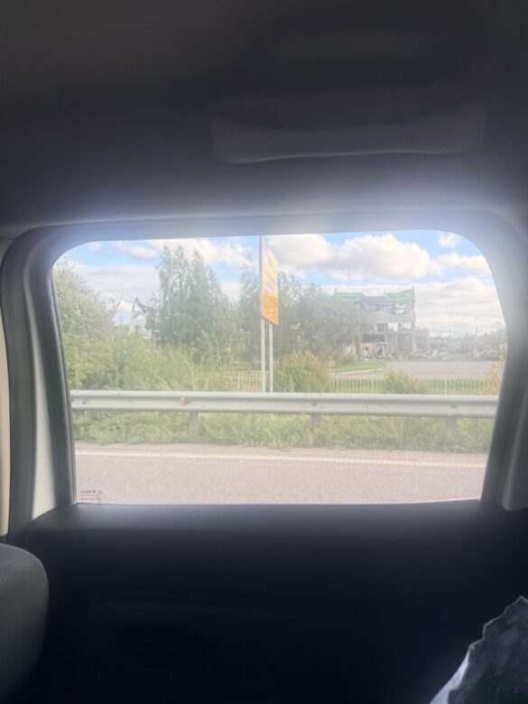 View from a car window: highway with a metal guardrail, green trees, and a distant construction site under a partly cloudy sky.