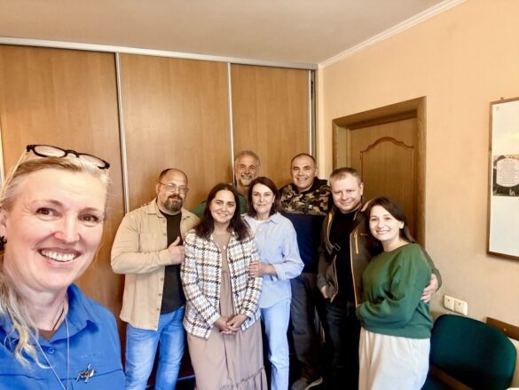 A woman in the foreground takes a selfie with seven people smiling behind her in a casual group photo indoors.