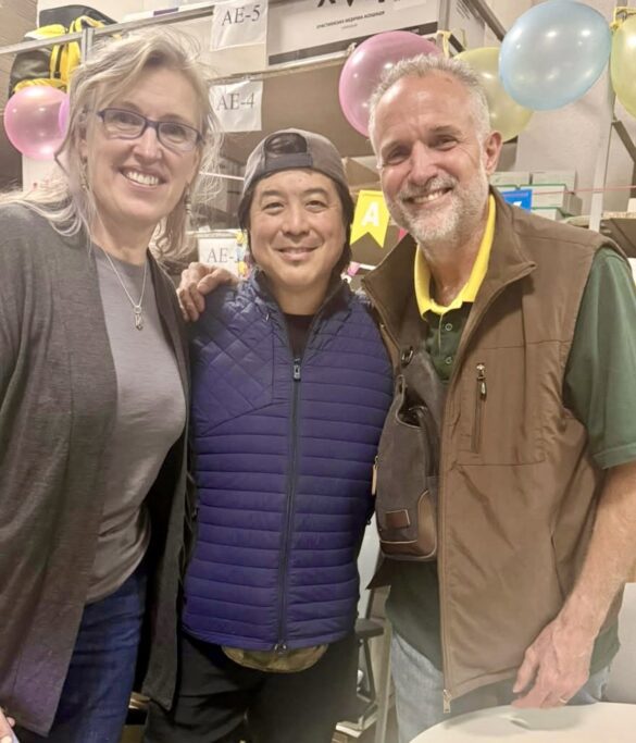 Three smiling adults posing together at an indoor event, with balloons and signage in the background.