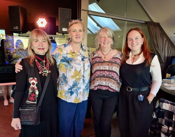 Four women pose together and smile for a photo at an indoor social event, standing side by side with arms around each other.