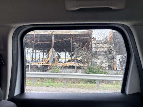 Burned-out industrial building with collapsed framework seen through a car window, rubble and charred remains exposed outside the road.