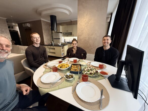 Four adults sit around a round white dining table in a modern kitchen, sharing a home‑cooked meal with bowls of salad and roasted potatoes, smiling at the camera, with a monitor visible on the right.