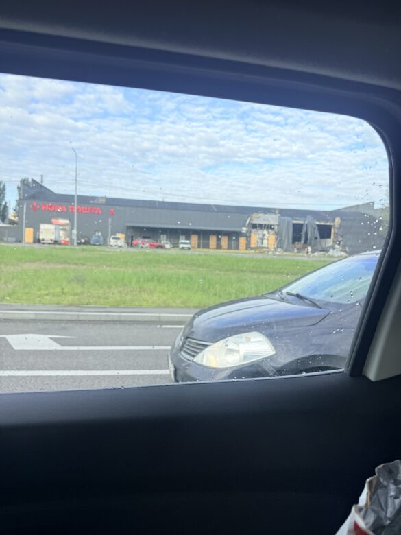 View from a car window of a building with a red Cyrillic sign (Nova Poshta) under construction, with parked cars outside and a grassy area nearby.