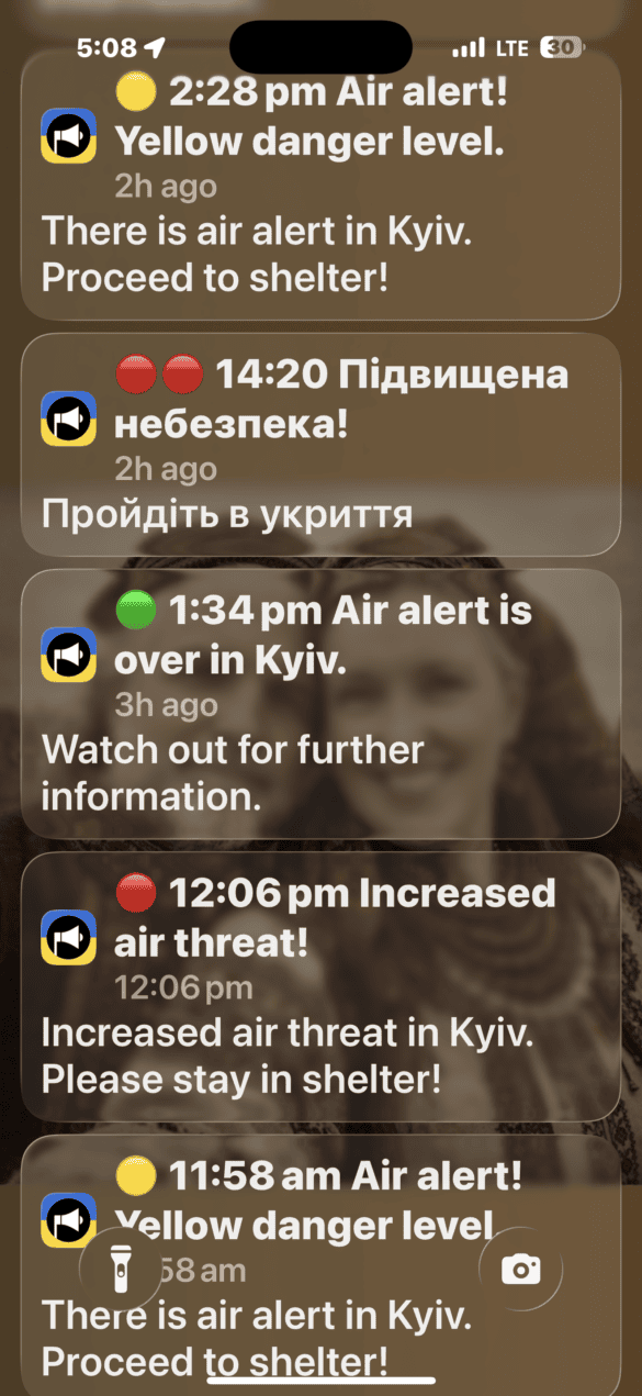 Stacked Kyiv air-alert notifications on a smartphone: rounded cards with times, color codes (yellow/red/green), Ukrainian text, and an app icon visible.