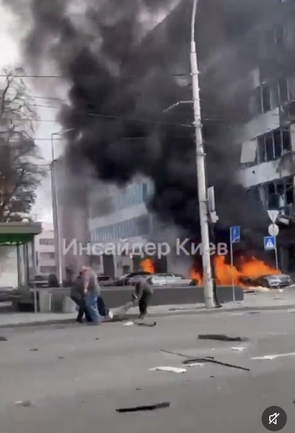 Street scene with black smoke and flames from burning cars while bystanders assist an injured person on the sidewalk/roadway edge.