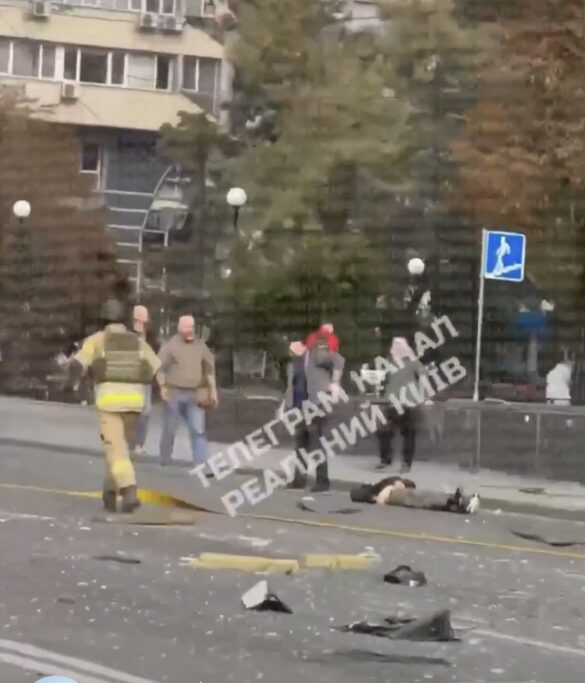 Firefighter in reflective gear approaching a person lying on a debris-strewn city street after an accident; several bystanders nearby.
