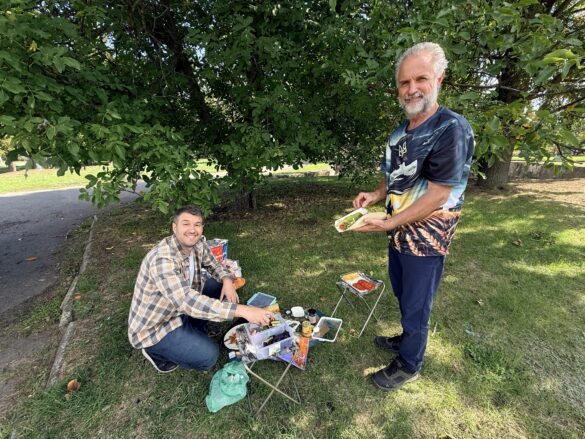Two men enjoy a park picnic under a leafy tree; one sits on the grass with a small cooler and food bags, the other stands holding a wrap plate.