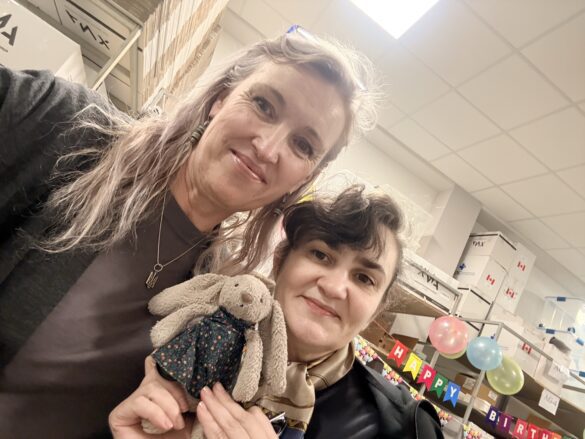 Two women pose for a selfie at a birthday party, one holding a plush bunny and a small girl’s toy, with balloons and a colorful banner in the background.