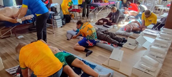 People lying on mats receiving first aid care in a medical training setup, with responders attending to them in a large room.