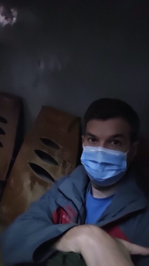 IMG_8330 Dimly lit room; man wearing a blue mask taking a selfie with stacked cardboard boxes in the background.
