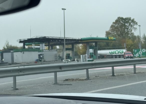 IMG_8480 Gas station with a WOG fuel tanker truck under a canopy, seen from the highway with a guardrail in the foreground.