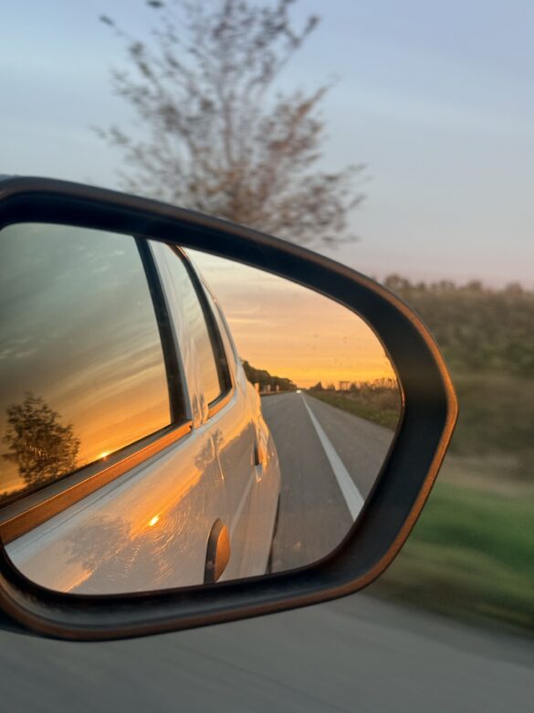 IMG_8518 Side mirror shows a sunset-lit road disappearing into the distance, with orange sky and a white car body reflected in the glass.