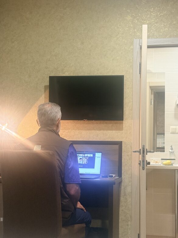 IMG_8542 Older man sitting at a desk in a hotel room, typing on a laptop with a blank TV on the wall above him.