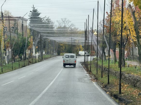IMG_8615 Street with a clear plastic net canopy stretched between tall poles over the road; a gray van travels away down the street.