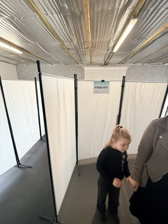 IMG_8657 Cardiologist screening area with white partition panels; child holding adult's hand walking through the space ( Ukrainian/Russian text 'КАРДІОЛОГ' on sign)