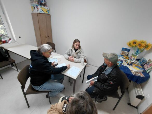 IMG_8701 Four people sit around a white rectangular table in a bright room, reviewing documents; a side table holds sunflowers and travel brochures at the back.
