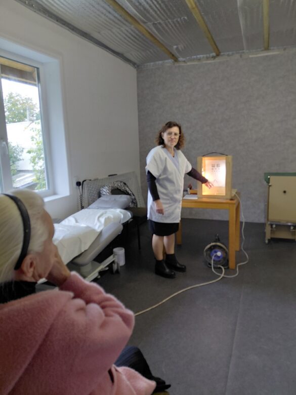 IMG_8703 Healthcare worker in a white coat demonstrates an eye-chart screening beside a table with a light box in a patient room.