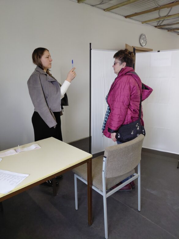 IMG_8705 A young woman in a gray jacket holds a blue pen toward an older woman in a pink puffer jacket at a check‑in area with privacy partitions.