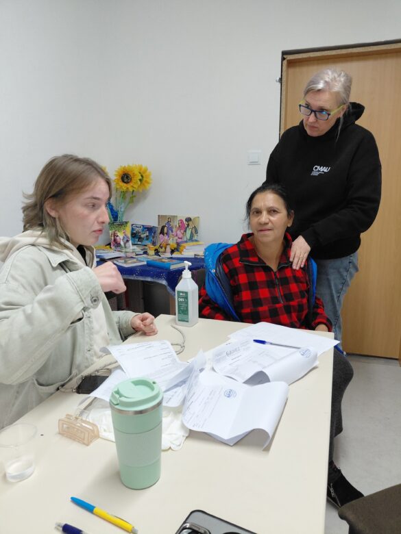 IMG_8710 Three people sit around a cluttered table with papers; a woman in a red plaid jacket sits while another stands beside her with a hand on her shoulder, suggesting guidance or support.
