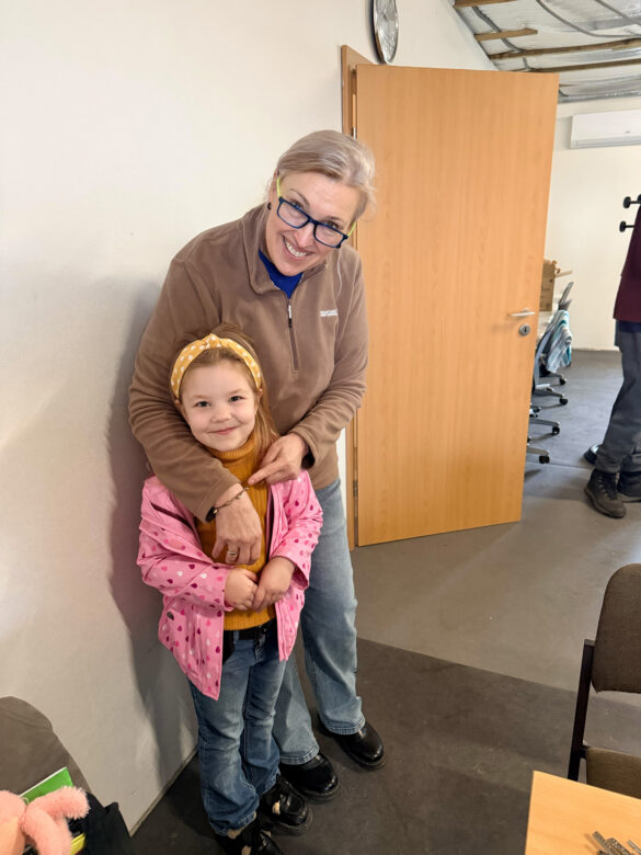 IMG_8743 Woman in a brown jacket hugs a smiling young girl in a pink polka-dot jacket indoors, by an open door.