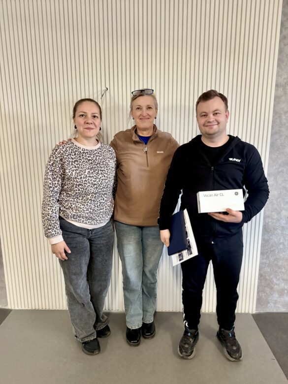 IMG_8744 Three adults posing indoors against a striped wall; the man on the right holds a box labeled Vscan Air CL.