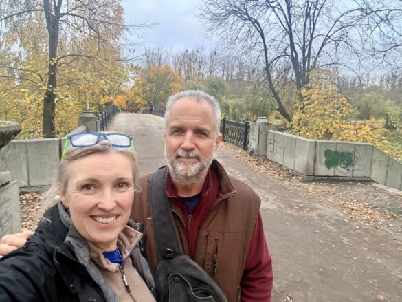 IMG_8775 Smiling couple taking a selfie on a stone bridge in an autumn park, with fallen leaves and colorful trees.