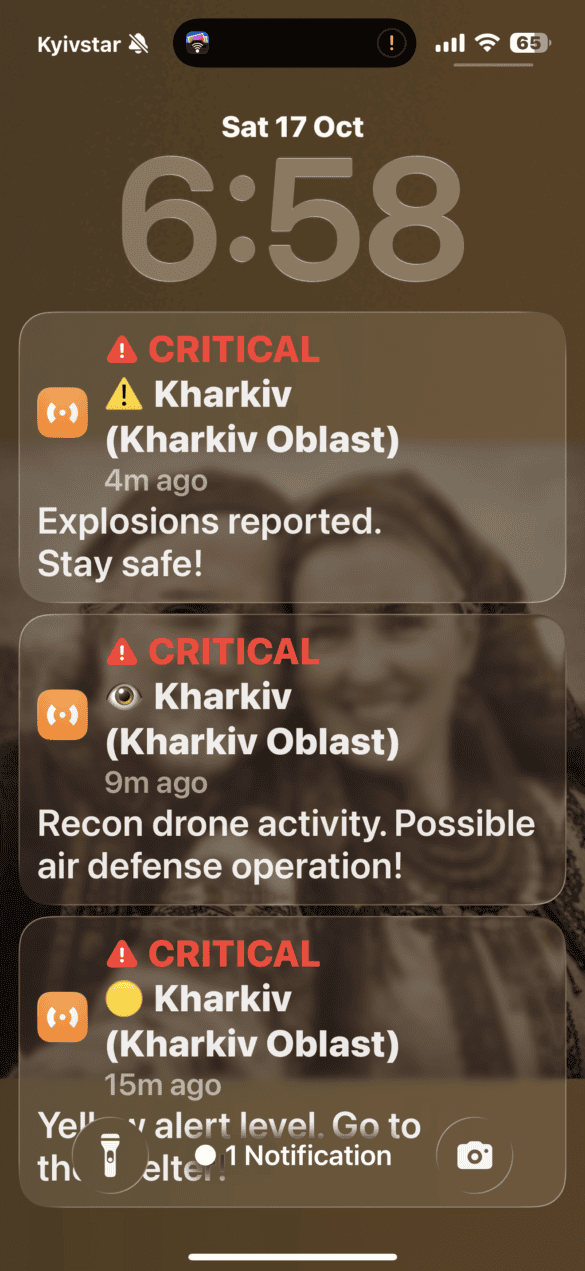 IMG_8822 Phone screen showing three critical Kharkiv alerts about explosions, drone activity, and yellow alert level, with a blurred background photo.
