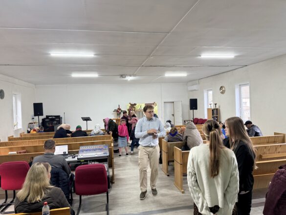 IMG_8857 People gathered in a hall with wooden pews, talking and standing; a man in a light shirt walks toward the camera as a stage with sound equipment sits in the back.