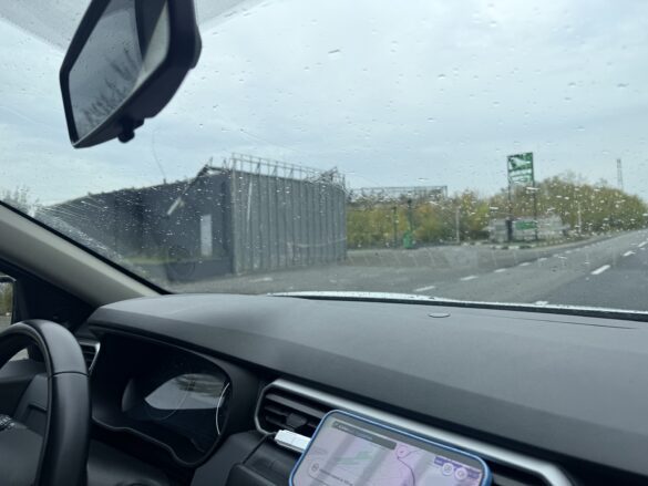 IMG_8869 Driver's view from a car on a rainy highway; raindrops on the windshield, a gray building on the left, and a green highway sign ahead.