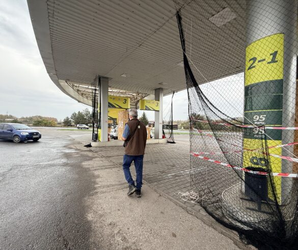 IMG_8873 Man in a brown vest and jeans walks toward fuel pumps at an outdoor gas station, with black safety nets and red tape nearby.
