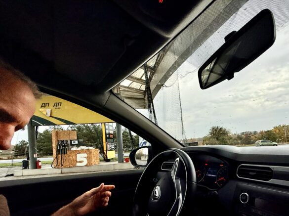 IMG_8874 Inside a car at a gas station, with the dashboard and steering wheel visible as a yellow canopy and fueling area appear outside through the window.